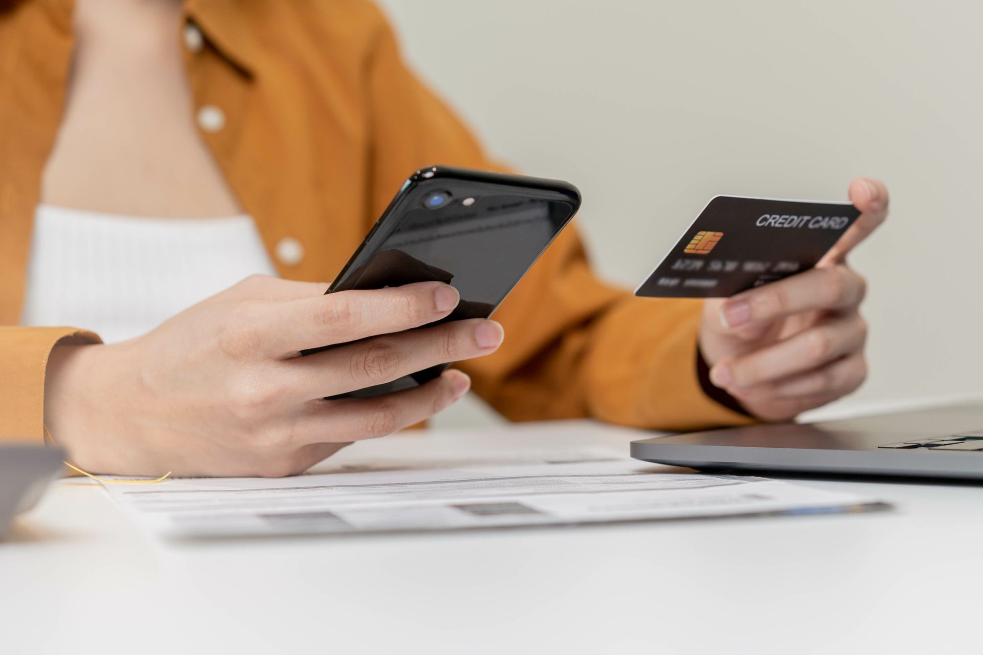 Foto zeigt eine Person vor dem Computer, die in einer Hand ein Smartphone und in der anderen eine Bankkarte h&auml;lt.