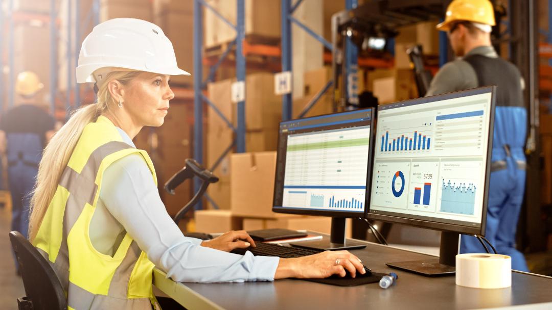 Foto einer Mitarbeiterin im Lager eines Unternehmens am Bildschirm, auf dem statistische Auswertungen einer Anwendung f&uuml;r Logistik und Lagerhaltung zu sehen sind