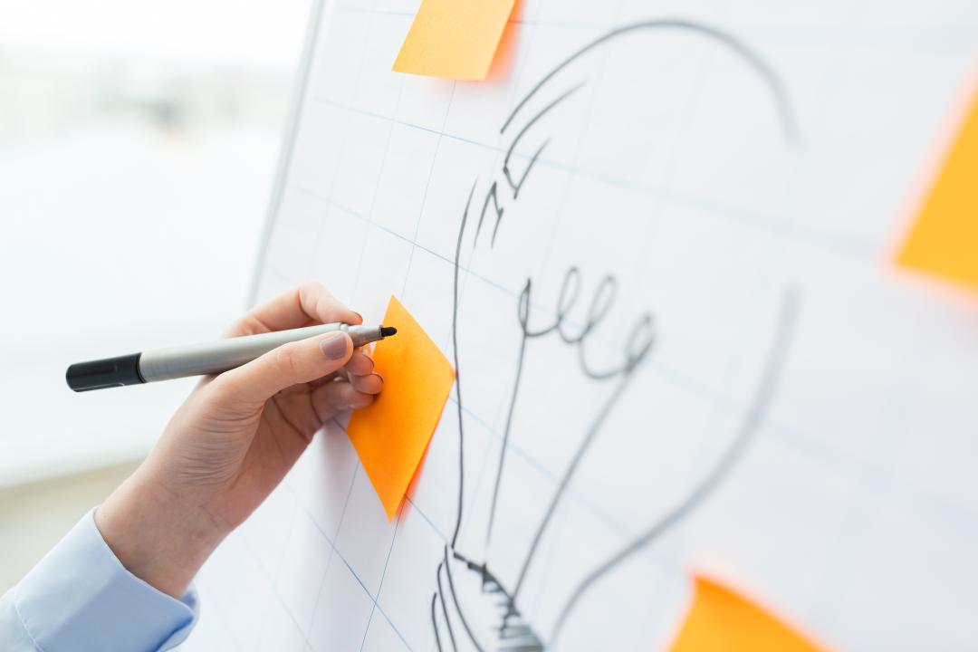 Foto eines Flipcharts, auf dem mehrere Post-its rund um die Skizze einer Gl&uuml;hbirne kleben, von denen eines aktuell von jemandem beschrieben wird.